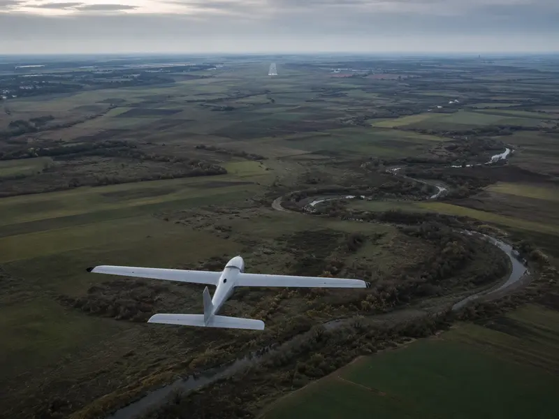 Pequeño dron de ala fija volando sobre campos hacia una pista situada a lo lejos